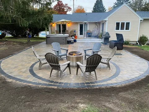 A landscaped backyard features a spacious stone patio behind a light-colored house with a covered porch. Outdoor seating surrounds a central fire pit, and warm lights illuminate the gathering area as evening approaches. Mature trees and neatly maintained landscaping create a cozy space for entertaining and relaxing outdoors.