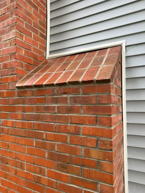 Close-up of a brick chimney with a vent or small window, next to a house with gray vinyl siding.