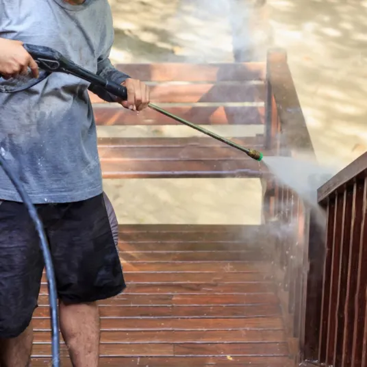 A person using a pressure washer to clean wooden stairs and railings outdoors, with water spraying and mist visible. The person is wearing a gray shirt and black shorts.