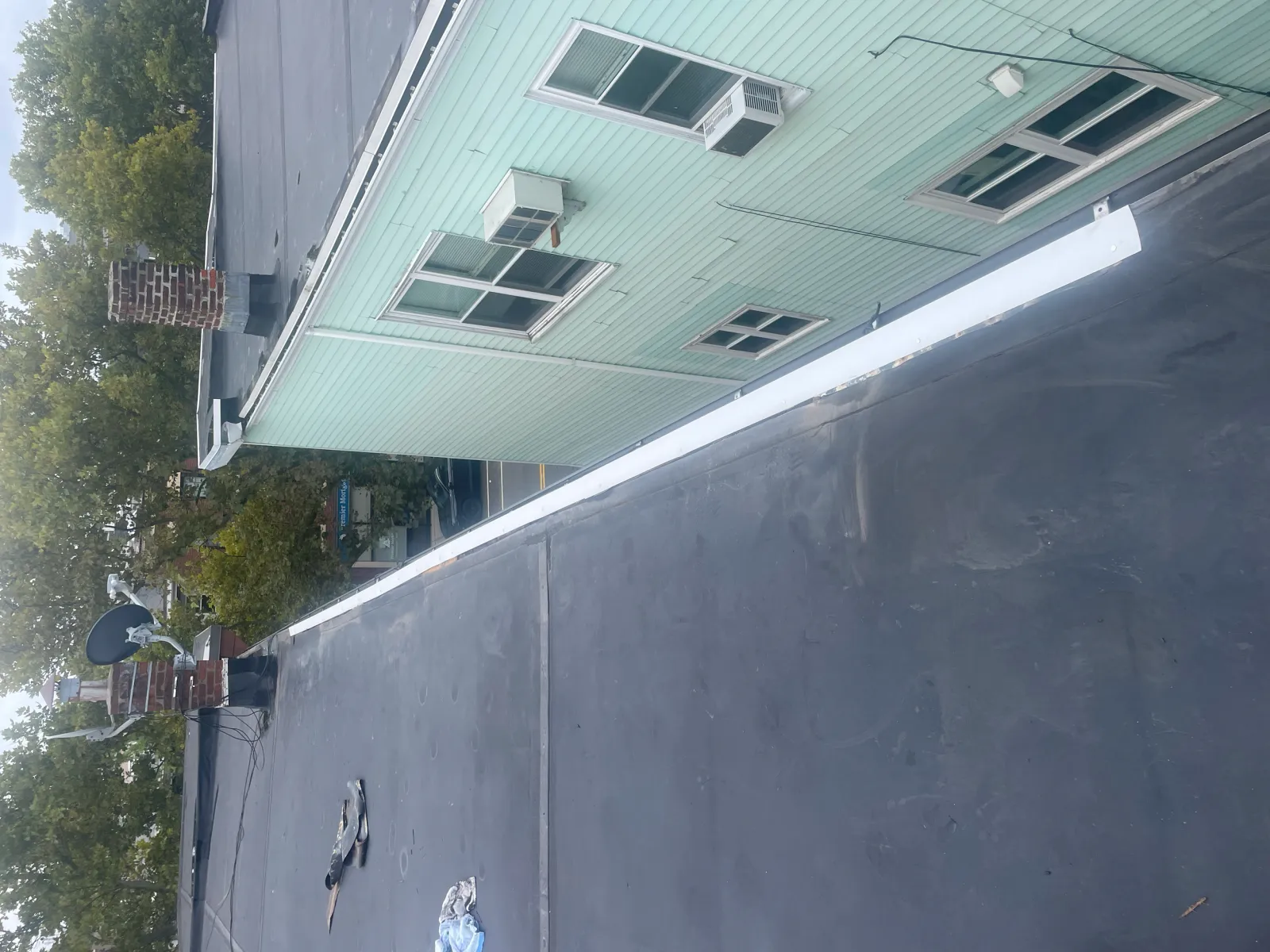 Flat rooftop with asphalt surface, chimney, satellite dish, and an adjacent mint green building with windows and air conditioners.