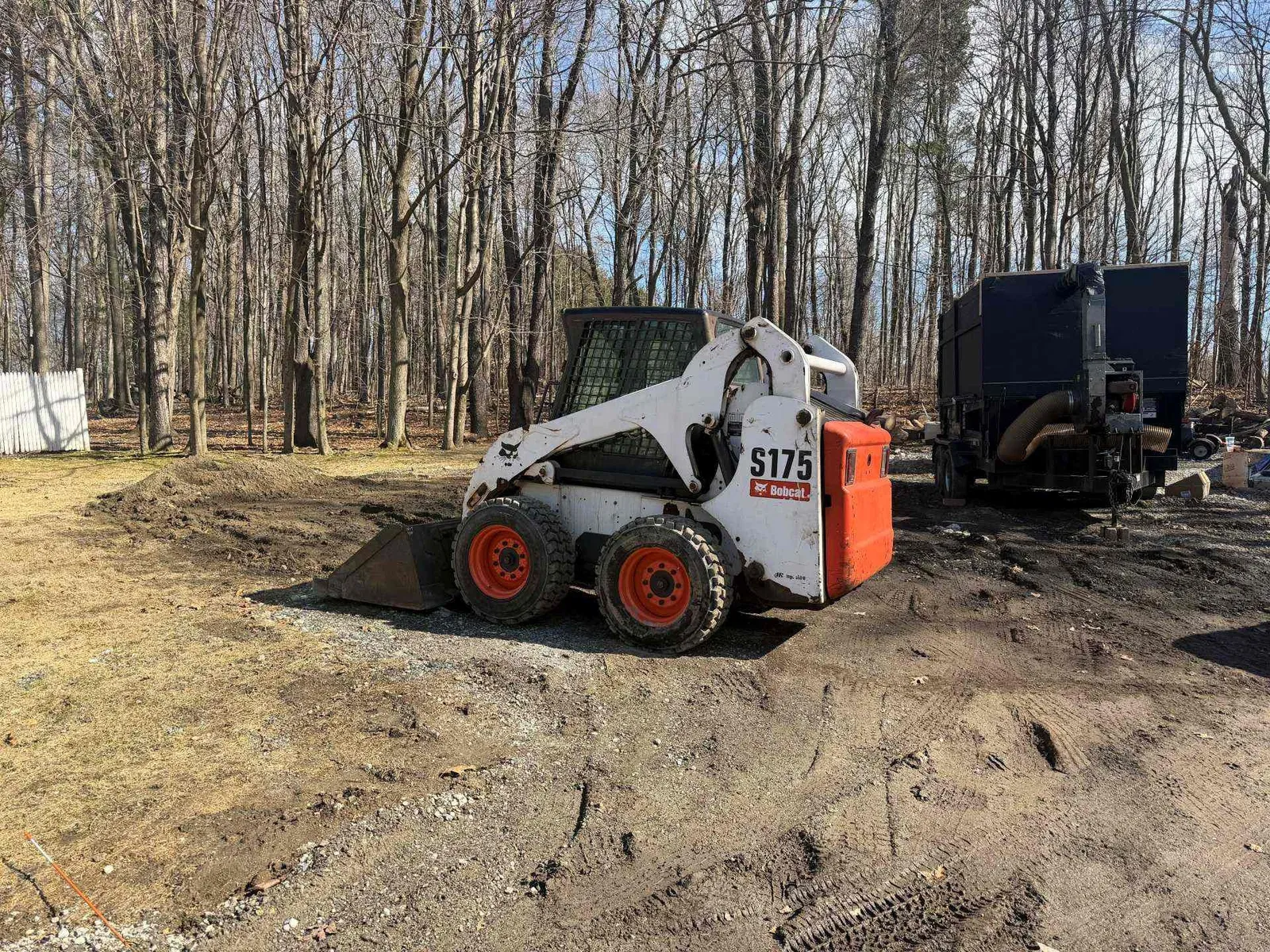 White skid steer loader with red wheels parked on a dirt lot near a tree line and trailer.