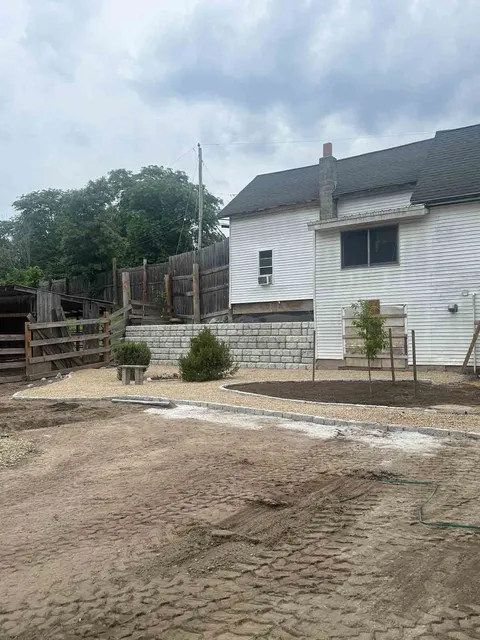 White house with a tiled roof beside a dirt yard and fence under a cloudy sky