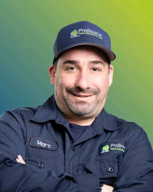 A smiling person wearing a dark blue button-down shirt with a name tag and a branded cap against a green background.