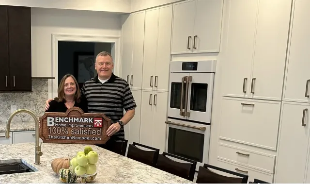 A homeowner couple holding a wooden Benchmark “100% Satisfied” sign in their finished white kitchen with granite counters