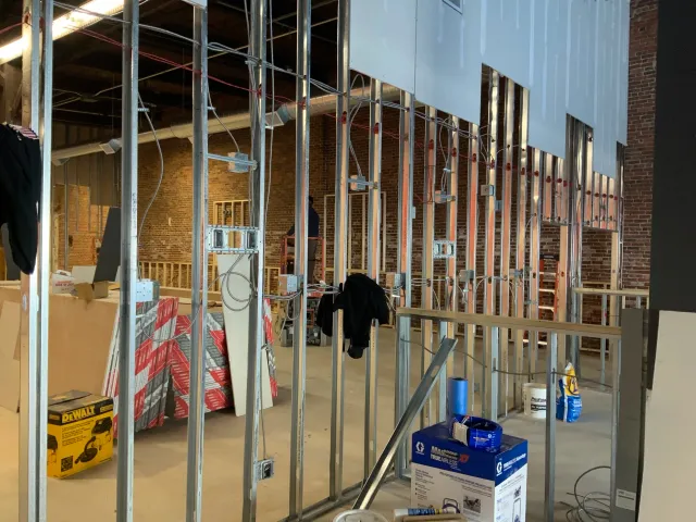 Interior construction site with exposed metal studs, wiring, drywall sheets, and construction materials. Brick wall is visible in the background.