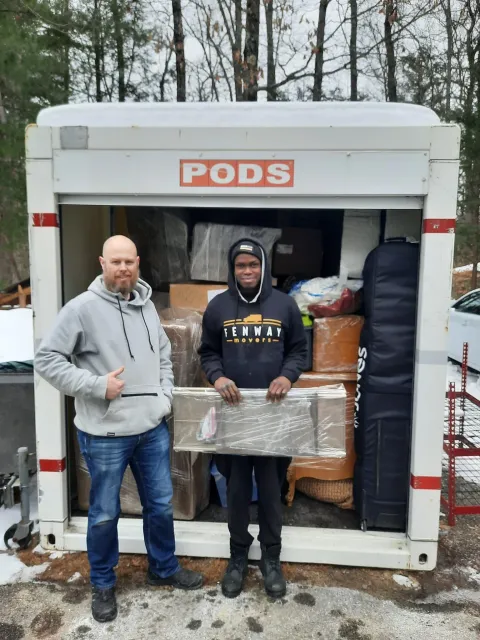 Two men are standing in front of a pods trailer holding boxes.