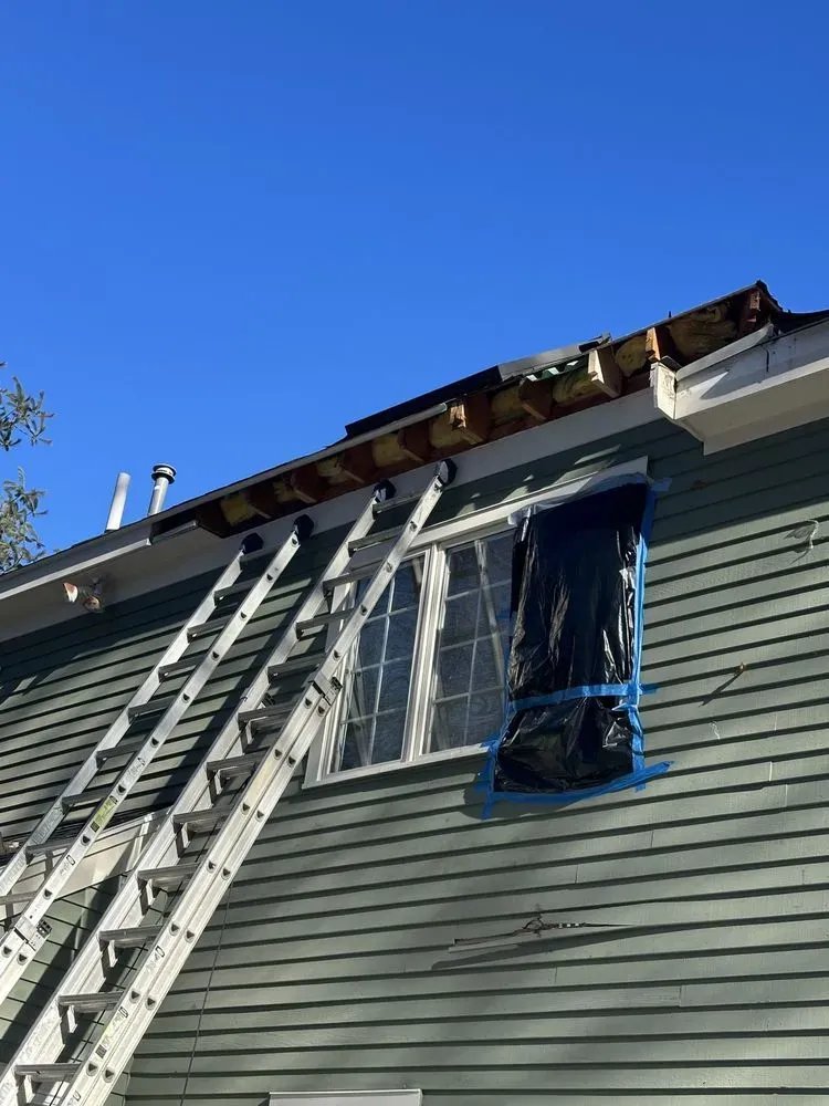 Ladders against a house with roof damage and a blue-taped window under a clear sky