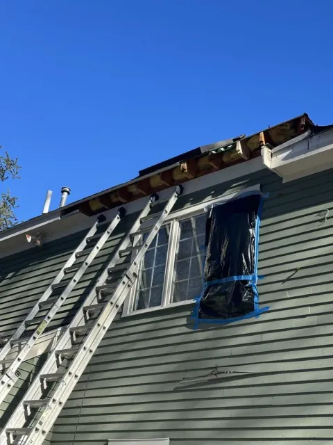 Ladders against a house with roof damage and a blue-taped window under a clear sky