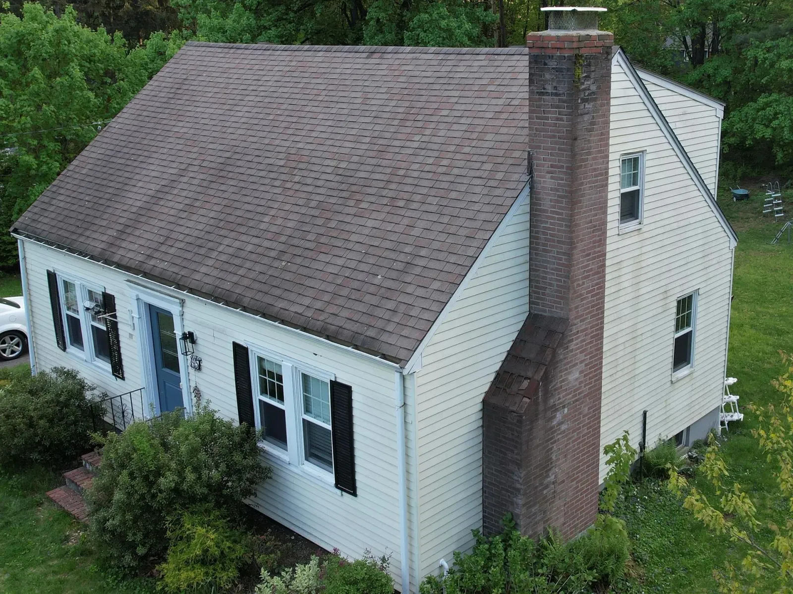 A white house with a steep shingled roof and brick chimney, surrounded by green trees and shrubs.