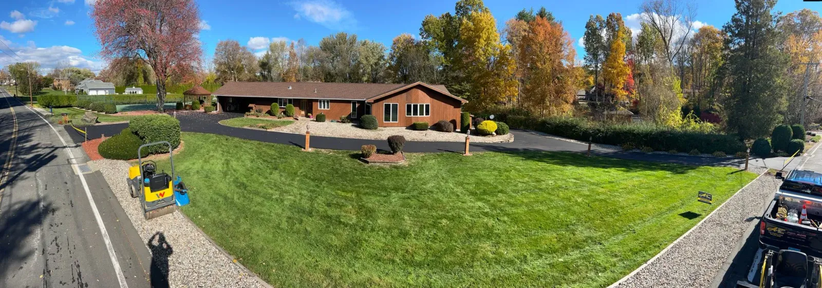 A panoramic view of a brown ranch-style house with an asphalt driveway and a grassy front lawn on a sunny autumn day.