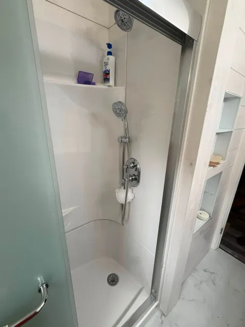 Walk-in shower with chrome showerhead, grab bar, shelf, and bottled toiletries.