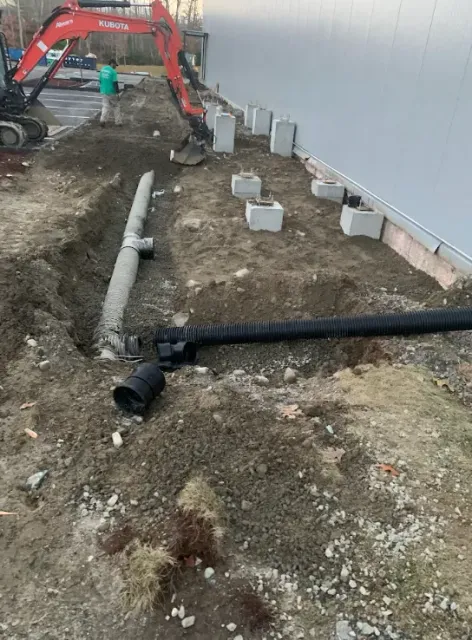 Construction site with excavator, workers, and pipes laid along a building wall under renovation