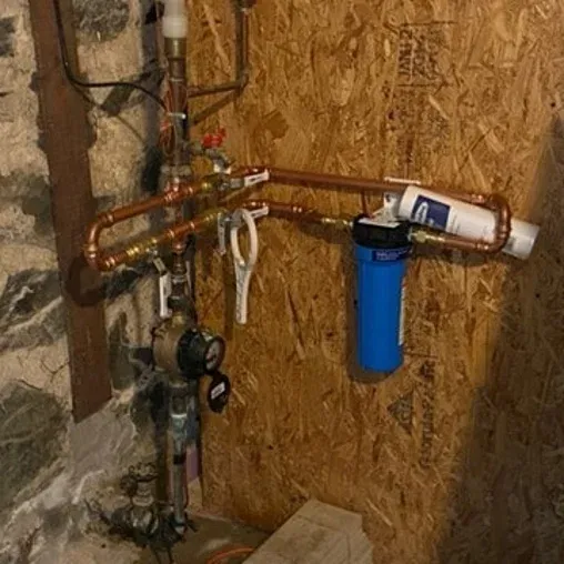 A home water filtration system with copper pipes and a blue cylindrical filter housing mounted on a plywood wall beside a stone foundation. Various valves, gauges, and fittings are attached to the plumbing.