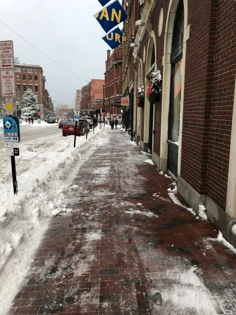 Snowy brick sidewalk next to a red brick building, street lined with snow-covered roads and buildings.