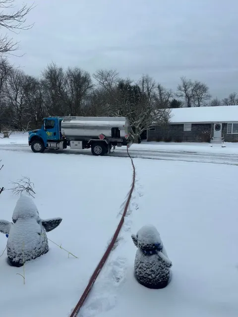 A blue fuel delivery truck is parked on a snowy road, with a hose running across the snow to a nearby house.