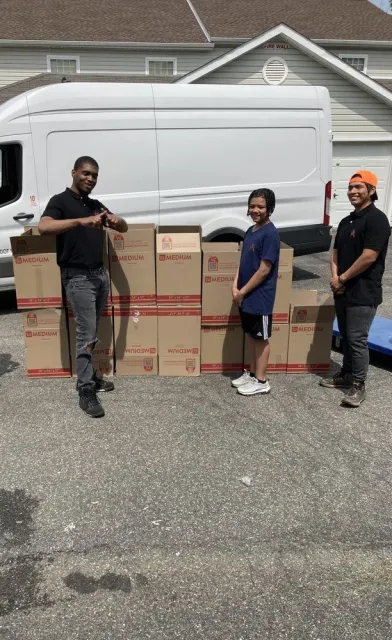 Four people standing with boxes in front of a white van on a driveway.