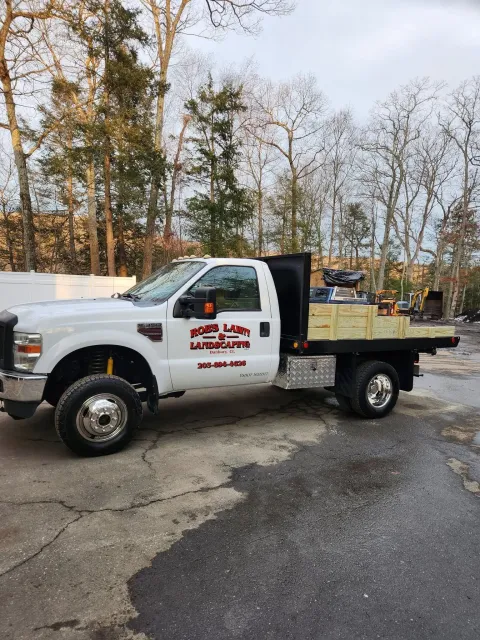 White utility pickup truck with company logo parked on a wet lot near trees and other work vehicles