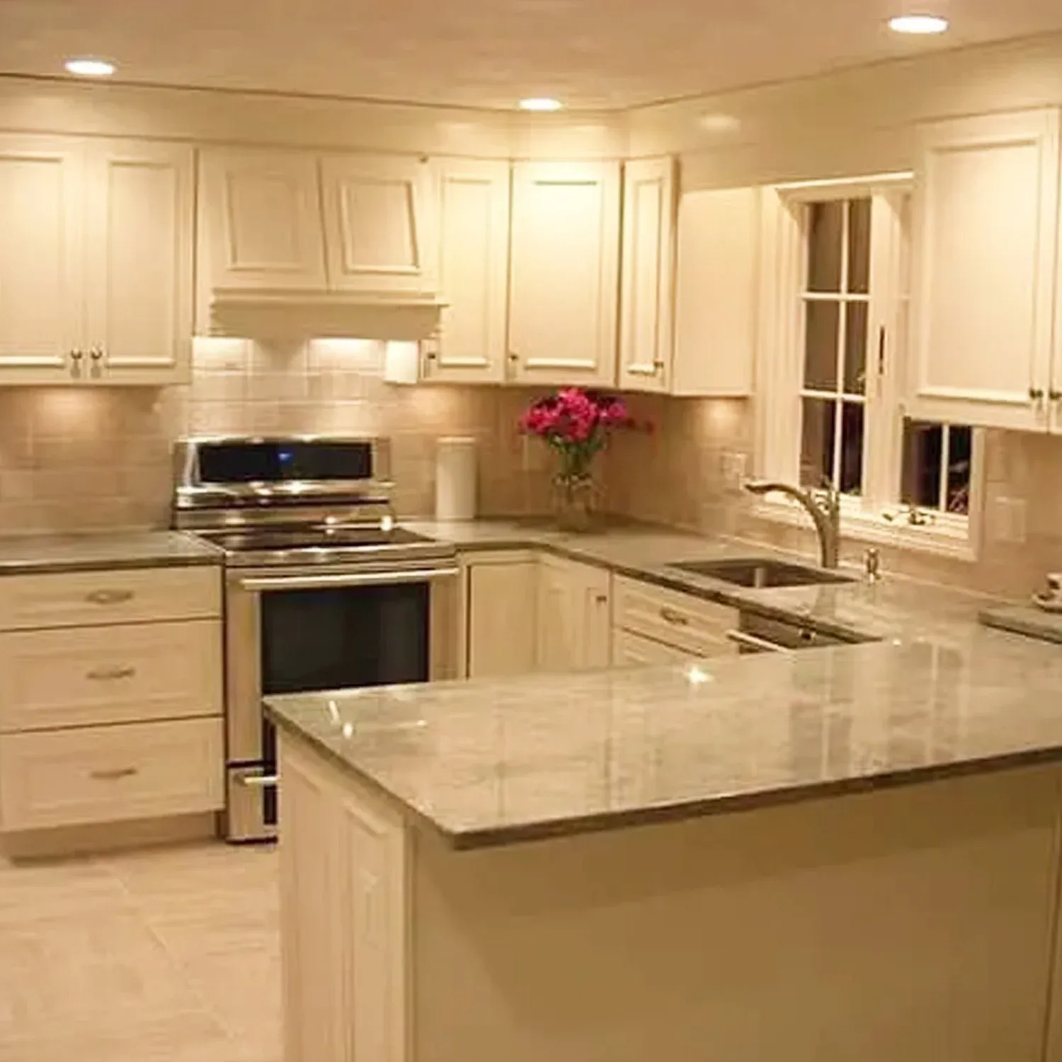 Kitchen with cream-colored cabinets, granite countertops, and a window above the sink. There is a black oven, a bouquet of pink flowers on the counter, and recessed ceiling lights.