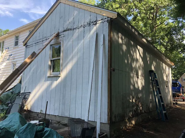 A side view of a white, vertical-sided shed under construction, showing a window, an attached ladder, and exposed wood.
