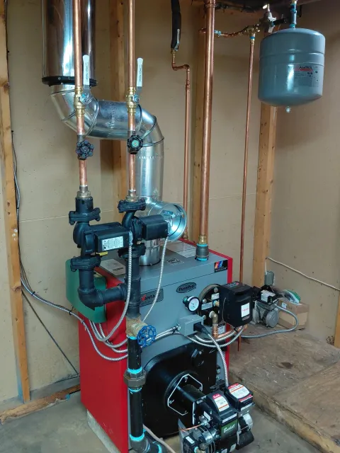 A boiler system with copper pipes, a pressure relief tank, a circulation pump, and various control valves installed on a concrete floor in a utility room.