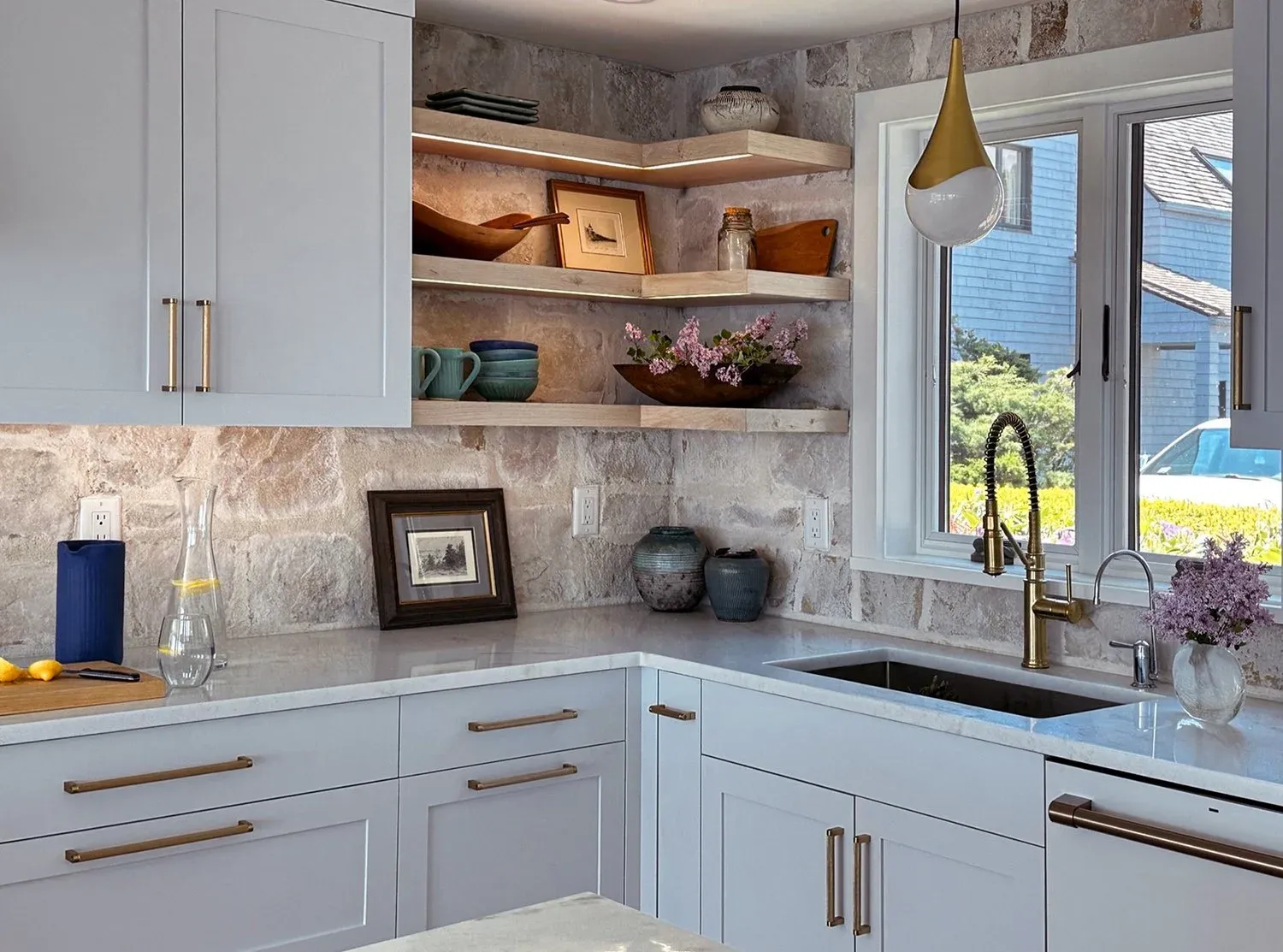 Modern kitchen with white cabinets, dramatic stone backsplash, marble countertop, open wood shelves, brass faucet, and a window view of the neighborhood.