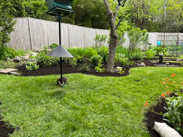 A well-maintained backyard garden with lush green grass, decorative plants, a tree, a white picket fence, and various garden decorations including a bird feeder, a solar light, and a blue wind spinner.