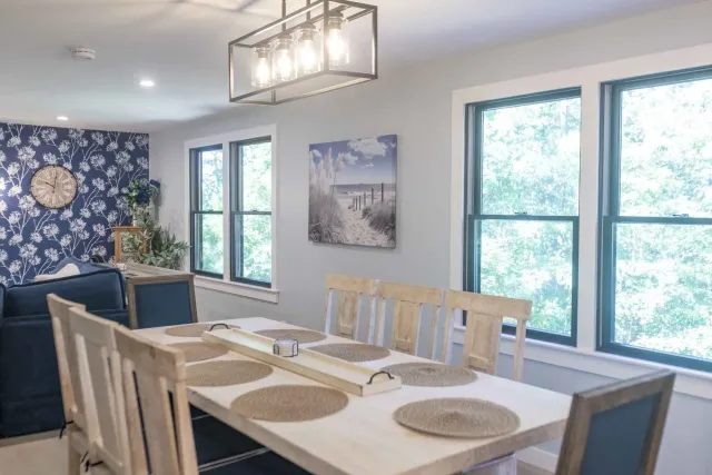 Bright dining room with wooden table, woven placemats, blue chairs, and large windows overlooking greenery