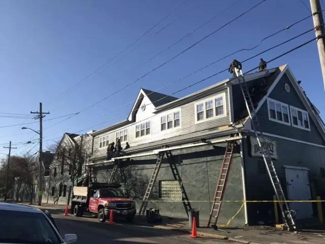 Workers on ladders repair the roof of a blue-gray building on a sunny day, with a red truck parked nearby.