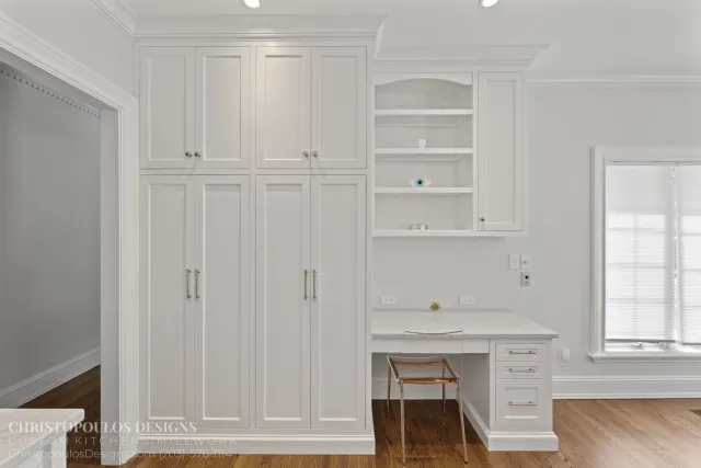 White kitchen cabinets with multiple doors and drawers, some open shelves, a small stool, and a window with blinds. The floor is hardwood, and there are electrical outlets and a partially visible countertop.