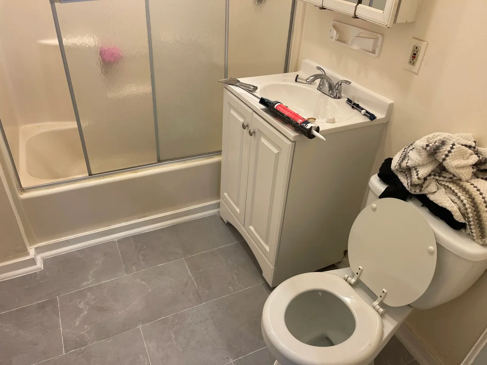 A small bathroom area with a shower stall, a white sink with a cabinet underneath, a toilet, and a pile of towels on top of the toilet tank. There are some tools and a caulking gun on the sink, indicating work is being done.