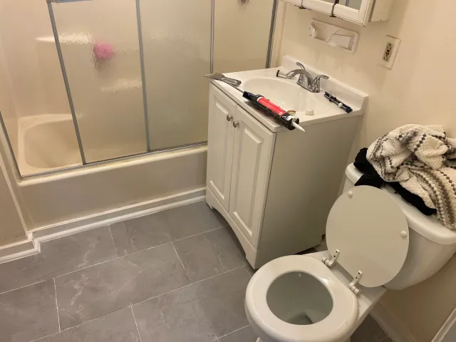 A small bathroom area with a shower stall, a white sink with a cabinet underneath, a toilet, and a pile of towels on top of the toilet tank. There are some tools and a caulking gun on the sink, indicating work is being done.