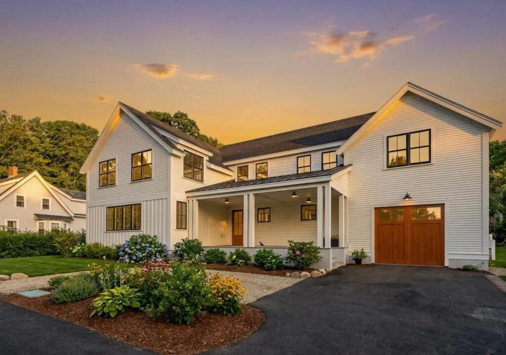 Modern two-story house with white siding, black-trimmed windows, wooden garage door, front porch, garden beds, and paved driveway at sunset.