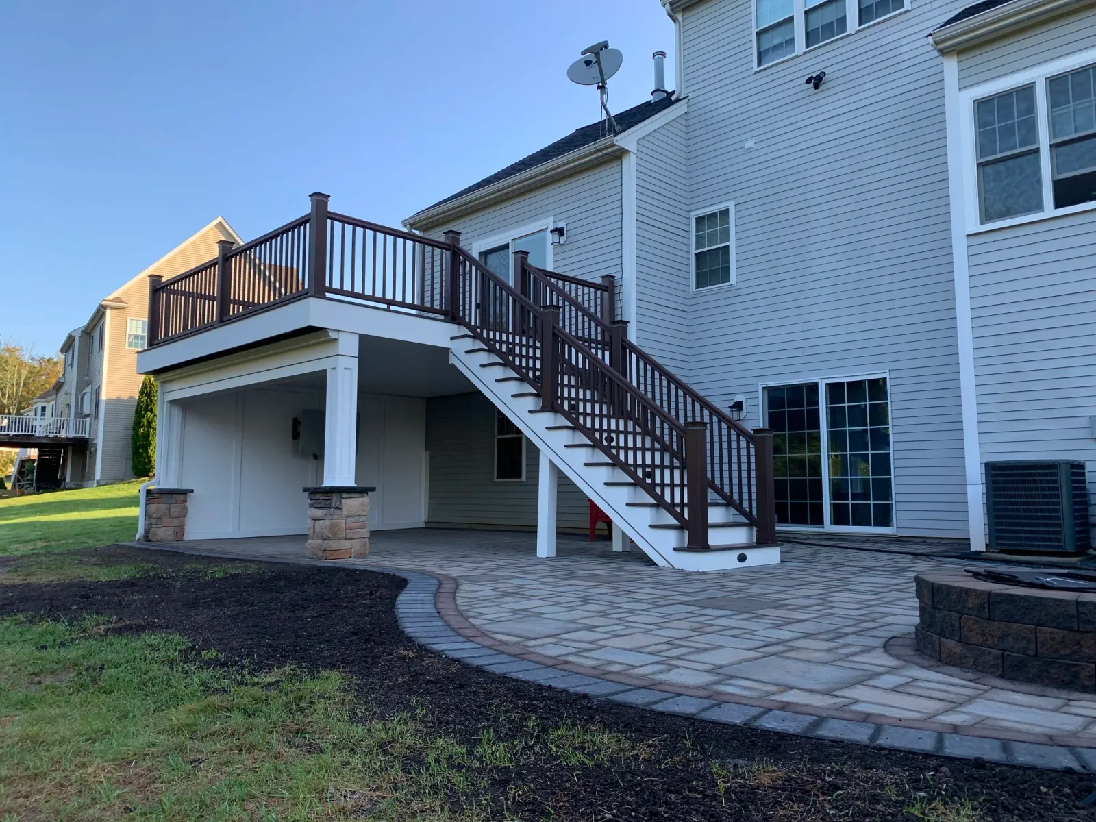 A backyard deck with stairs leading down to a stone patio, attached to a light-colored house, with a small grassy area in the foreground.