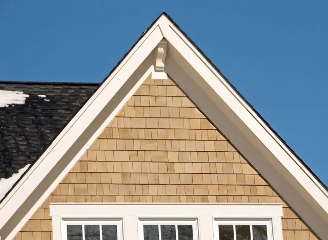 Close-up of a triangular house gable with beige wood shingles, white trim, and black roof tiles with some snow against a clear blue sky.