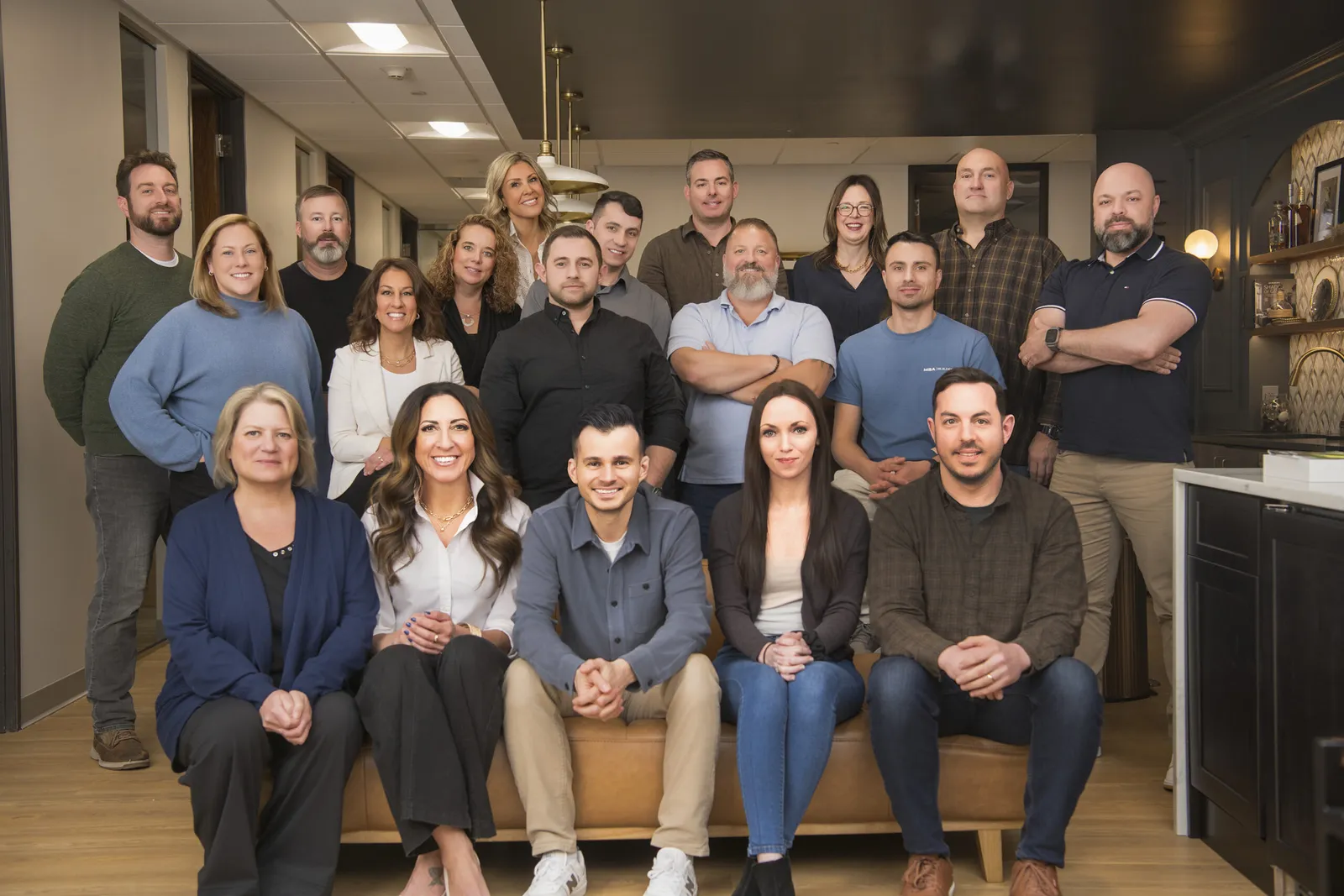 A group of seventeen adults pose together indoors, some seated on a brown couch and others standing behind, in a modern office setting—capturing the collaborative spirit of About MBA Builders.