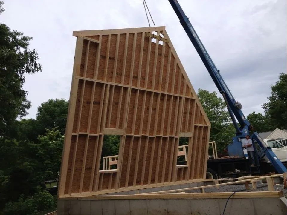 Framing of a house under construction being lifted by a crane, with trees and a cloudy sky in the background.