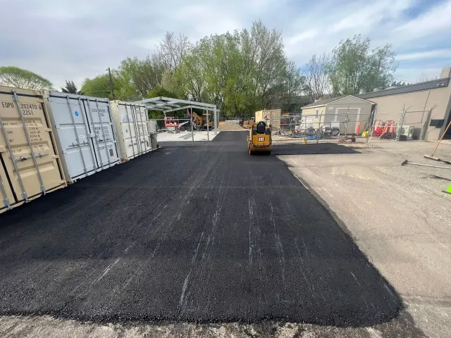 Black asphalt being laid. A yellow roller compacts the new pavement. Containers and building are visible.
