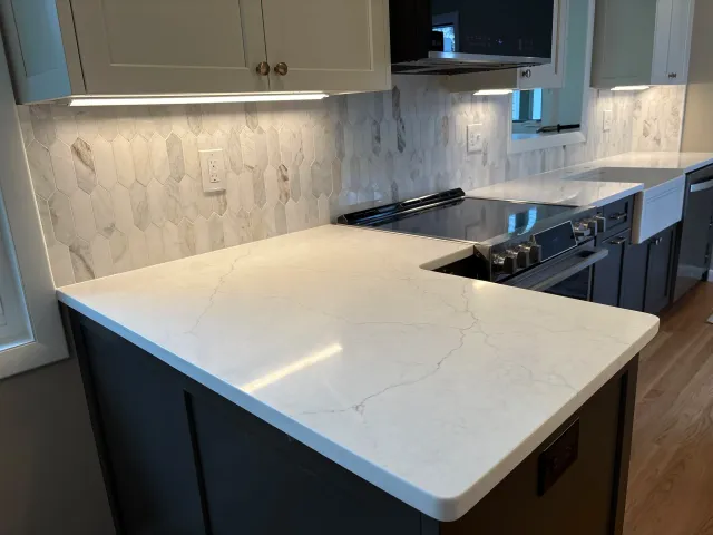 Kitchen with light-colored hexagonal tile backsplash, white marble countertop, and light gray cabinets with brass knobs. A window above the sink and a microwave are visible.