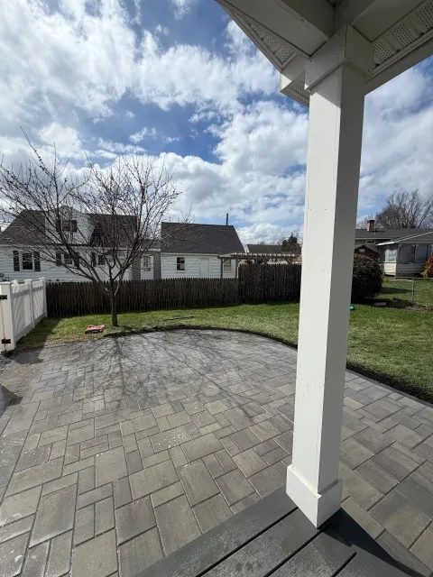 Covered backyard patio with gray pavers, lawn, fence, and cloudy sky.
