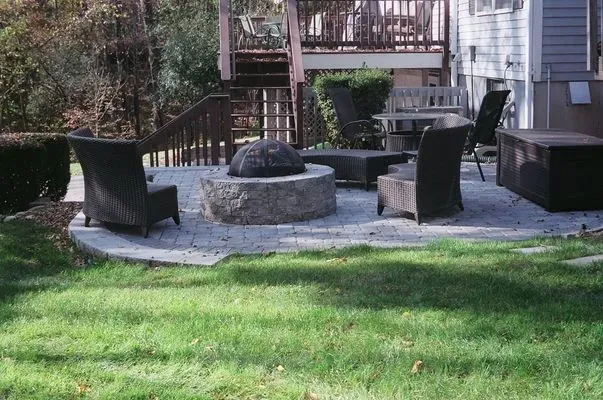 Circular patio with fire pit, chairs, and lawn, near a house and steps.