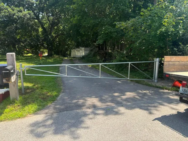 A metal gate blocks a paved driveway. A mailbox and trees are in the background.