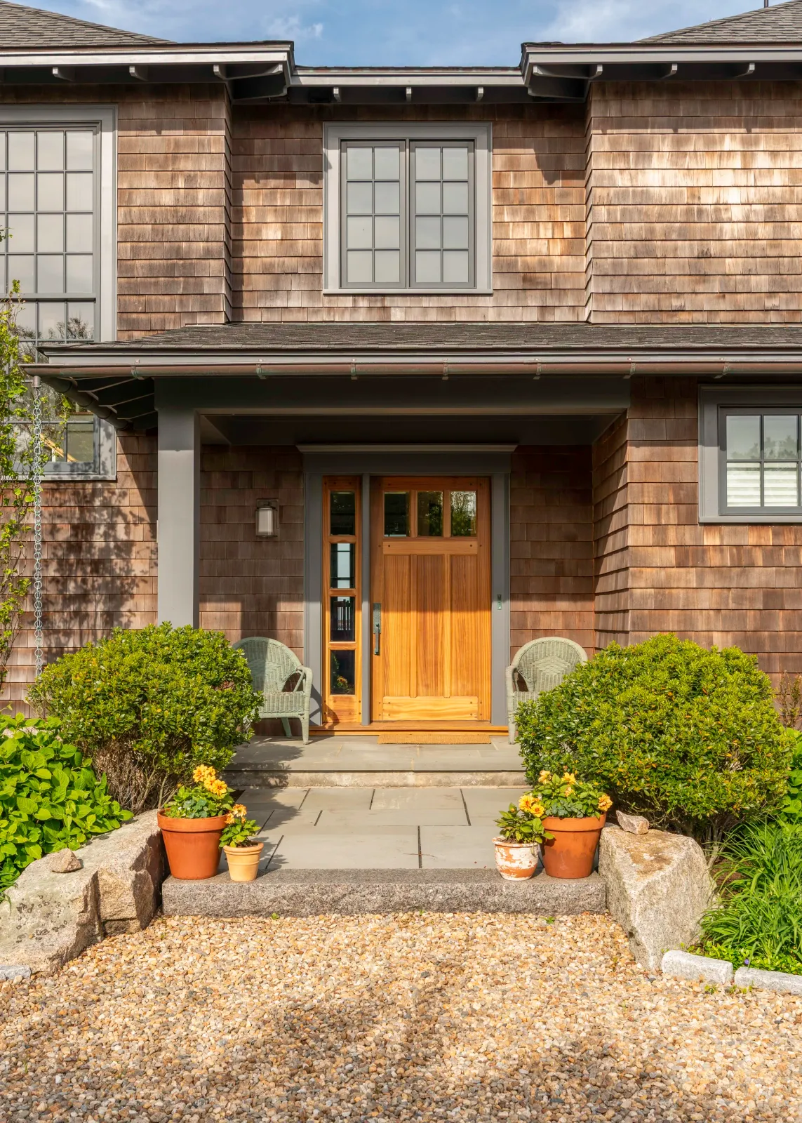 Sunny front entry of a traditional shingle-style home with a wooden door, two chairs, potted plants, and a gravel walkway.