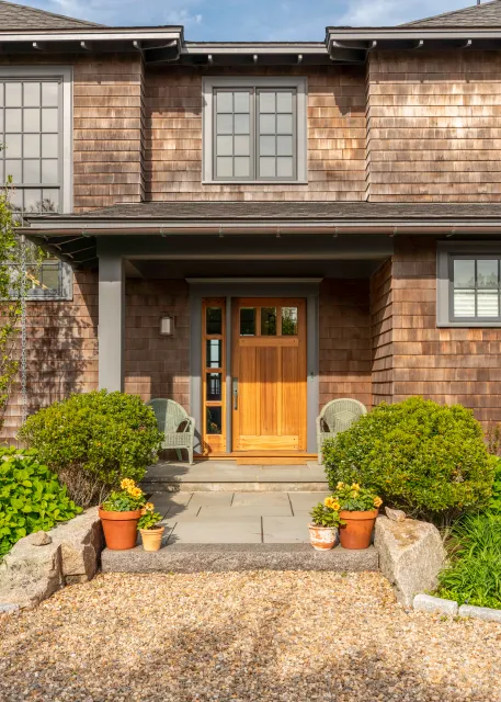 Sunny front entry of a traditional shingle-style home with a wooden door, two chairs, potted plants, and a gravel walkway.