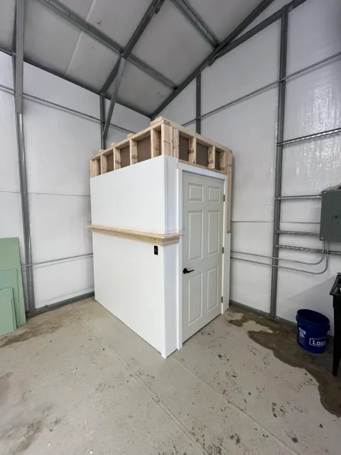 Interior of a building under construction with a white partition wall, a door, and exposed metal framing and insulation. There is a blue Lowe's bucket on the floor.