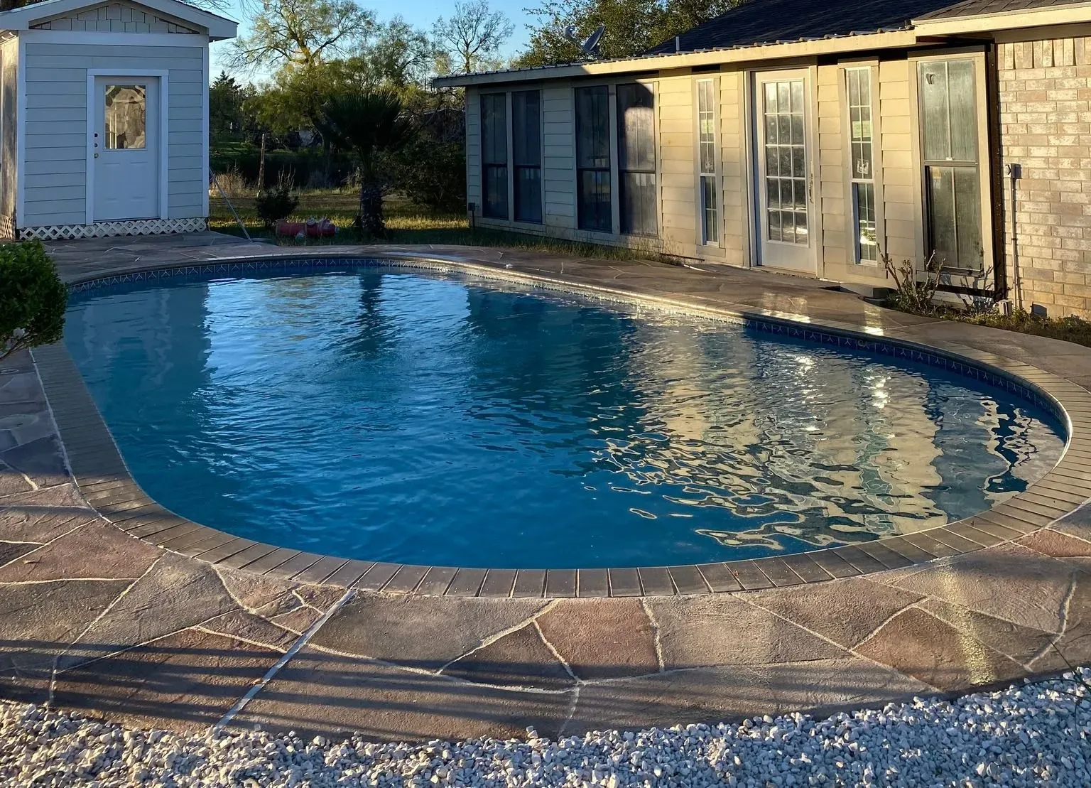 Oval-shaped swimming pool with stone edging, next to a light-colored building and shed. Blue water reflects the sky.