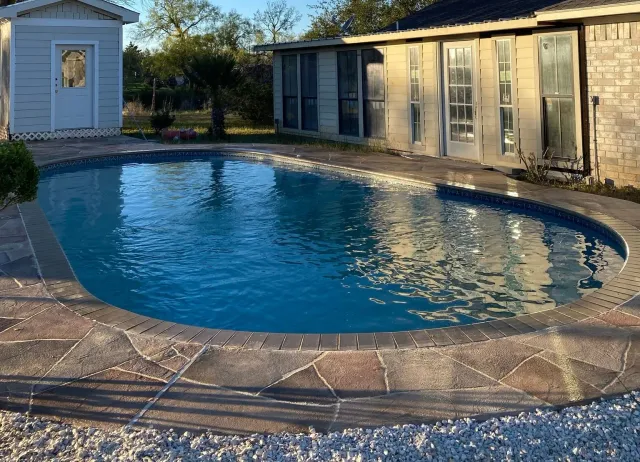 Oval-shaped swimming pool with stone edging, next to a light-colored building and shed. Blue water reflects the sky.