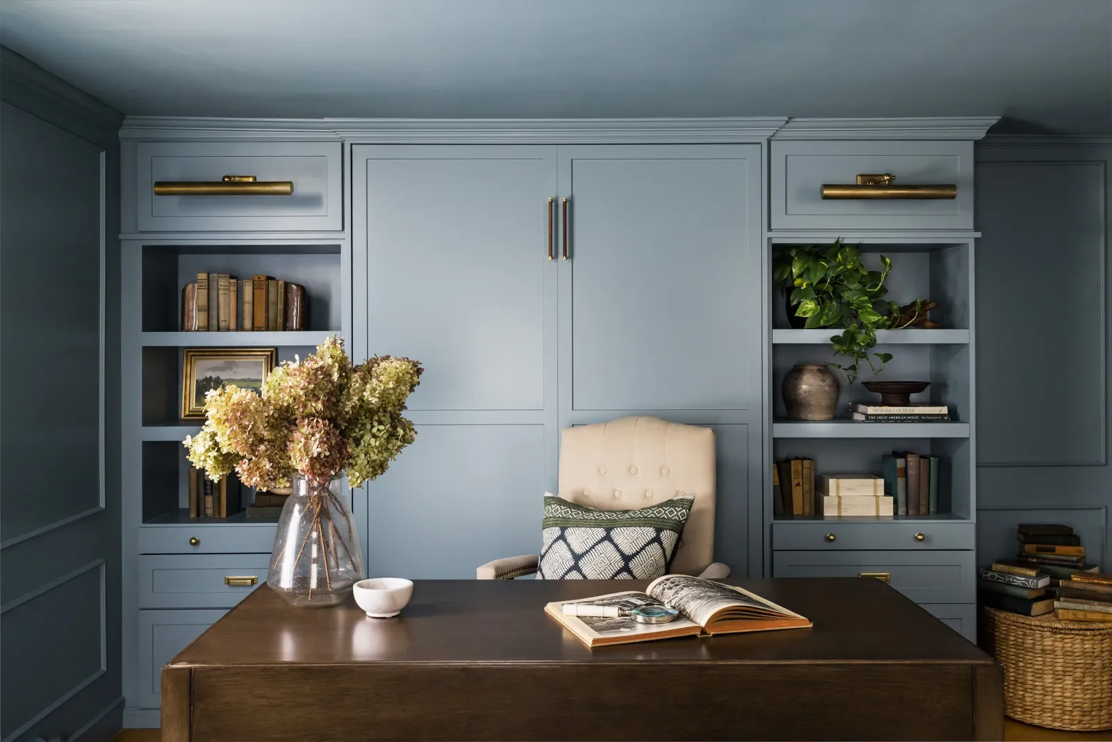 Elegant home office with blue built-in shelves, a beige upholstered chair, and a dark wooden desk. The shelves are filled with books, plants, and decor items. A vase with dried flowers is on the desk alongside an open book and a small bowl. Built in