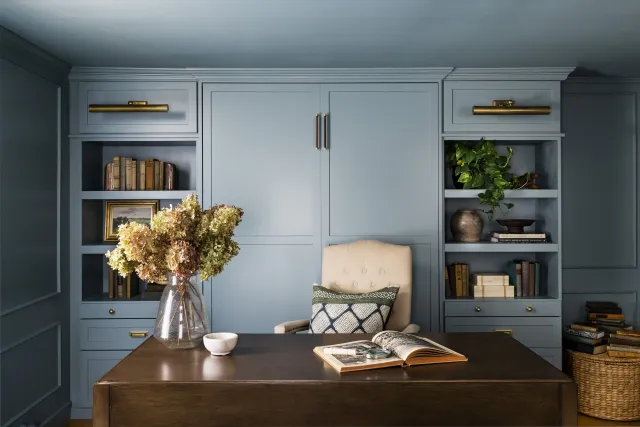 Elegant home office with blue built-in shelves, a beige upholstered chair, and a dark wooden desk. The shelves are filled with books, plants, and decor items. A vase with dried flowers is on the desk alongside an open book and a small bowl. Built in