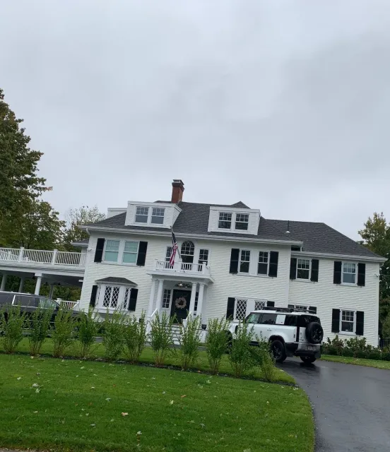 Large white colonial house with black shutters, American flag, and white SUV parked in the driveway