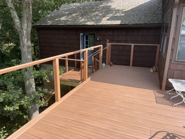 Newly built wooden deck outside a house, with a step leading to a backyard area, surrounded by trees and next to a brown house with a shingled roof.